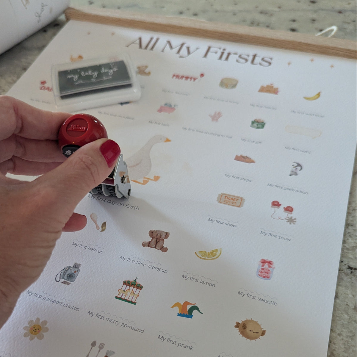 Hand holding a 'All My Firsts' Baby Goose edition milestone print on a countertop.